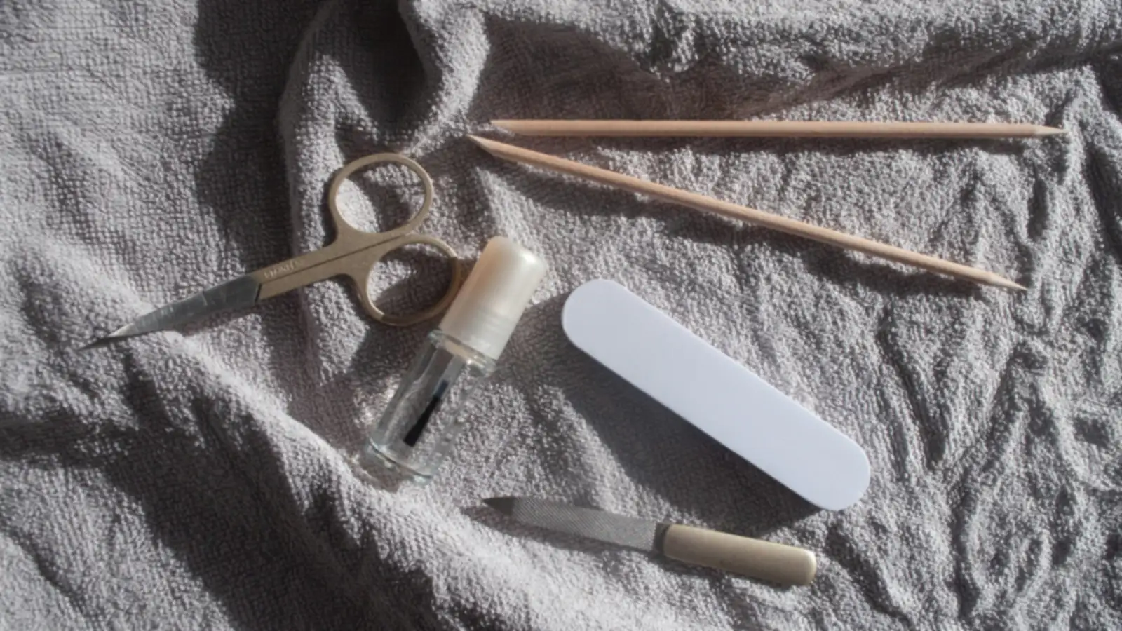 Manicure scissors, a nail file, a buffer and wooden sticks laid out on a gray towel