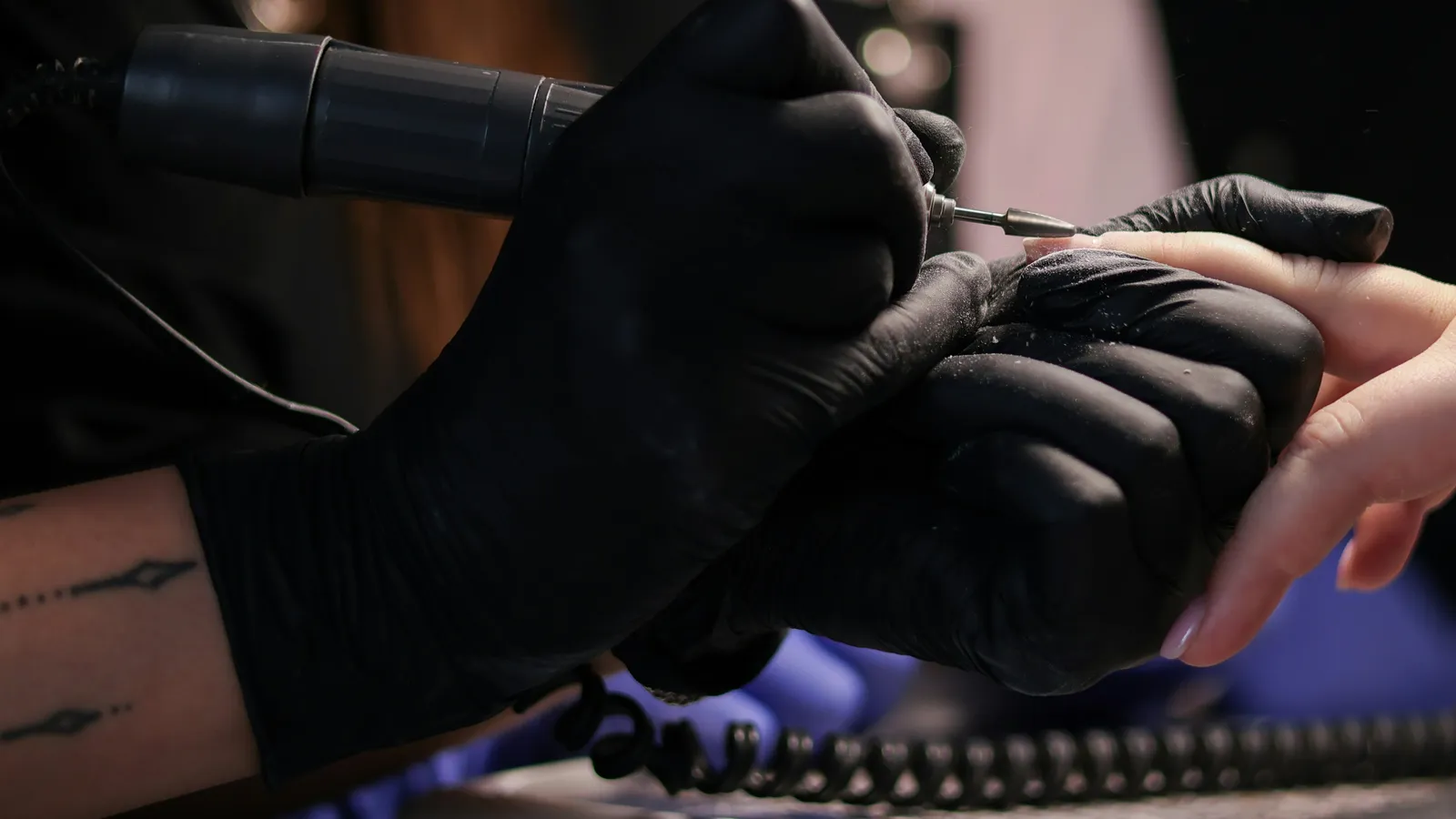 Gloved hands shaping a client's fingernail with an electric nail file